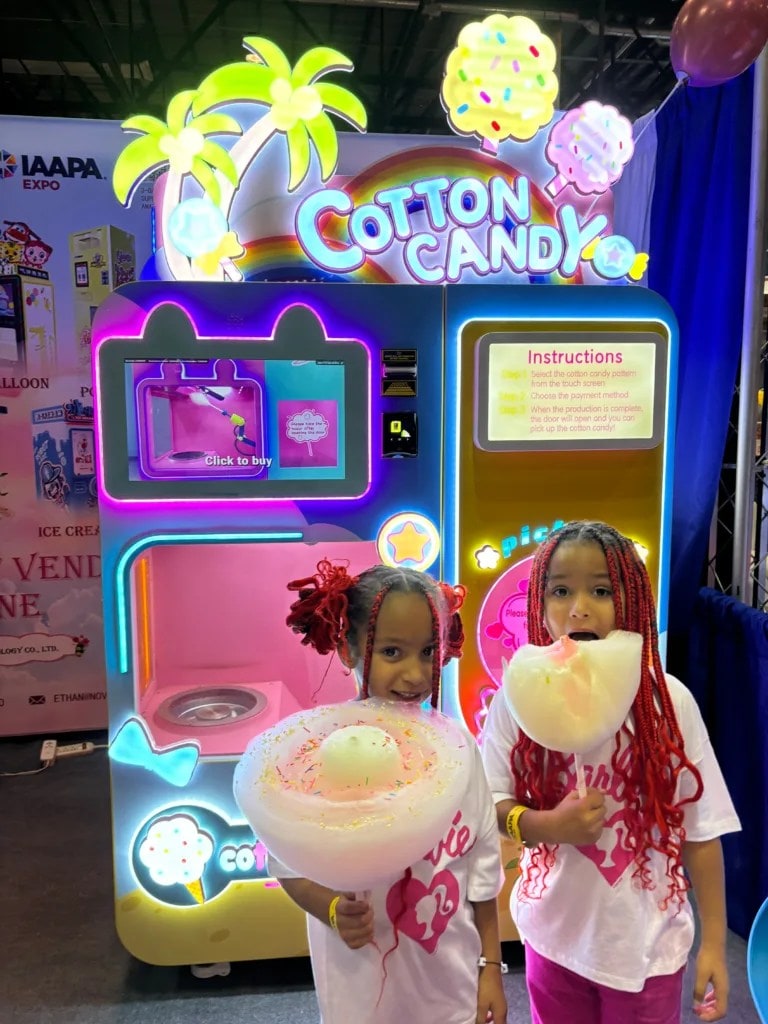 Two children holding cotton candy in front of a brightly lit cotton candy vending machine.