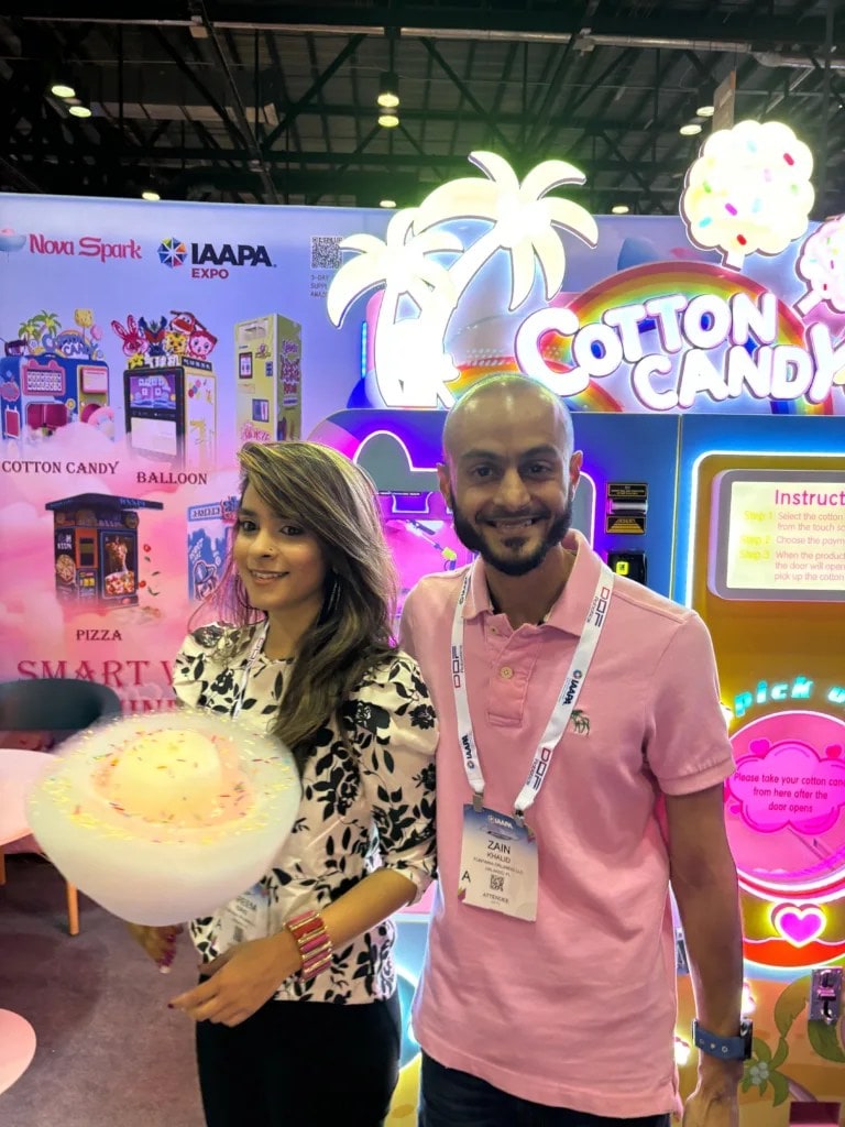 Two adults beside a cotton candy vending machine, one holding a flower-shaped cotton candy.