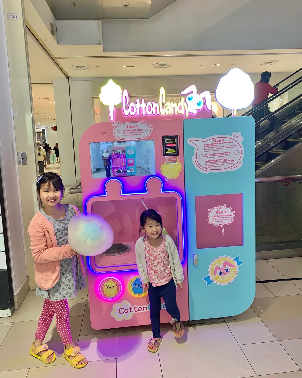 Two children beside a pink-and-blue cotton candy vending machine near an escalator.