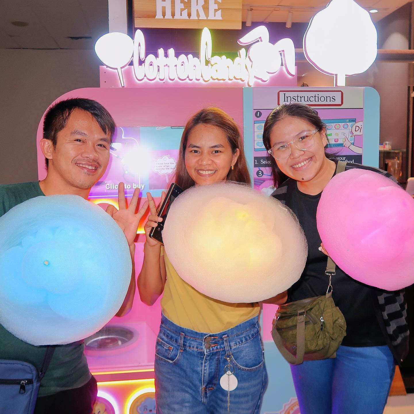 Three adults holding blue, yellow and pink cotton candy in front of a vending machine.