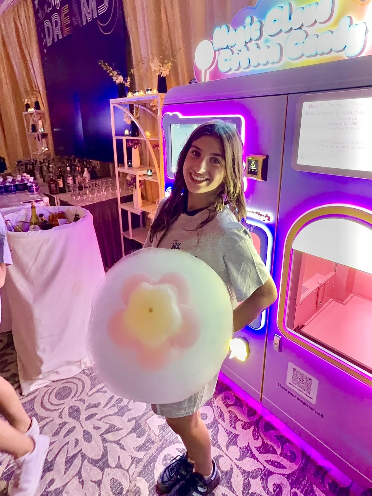 A woman holding a flower-shaped cotton candy beside a purple vending machine.