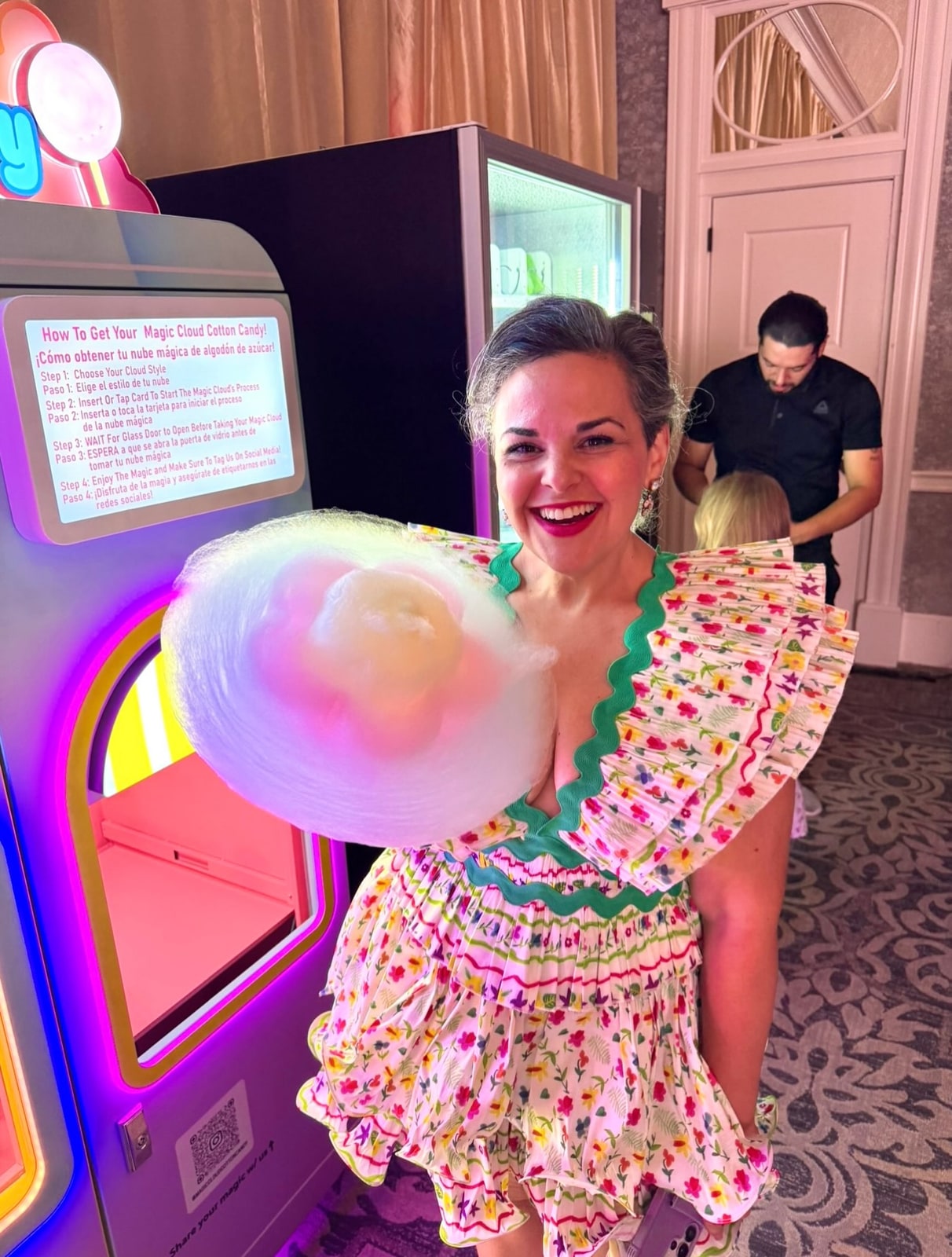 A woman in a floral dress holding a multicolored cotton candy beside the machine.