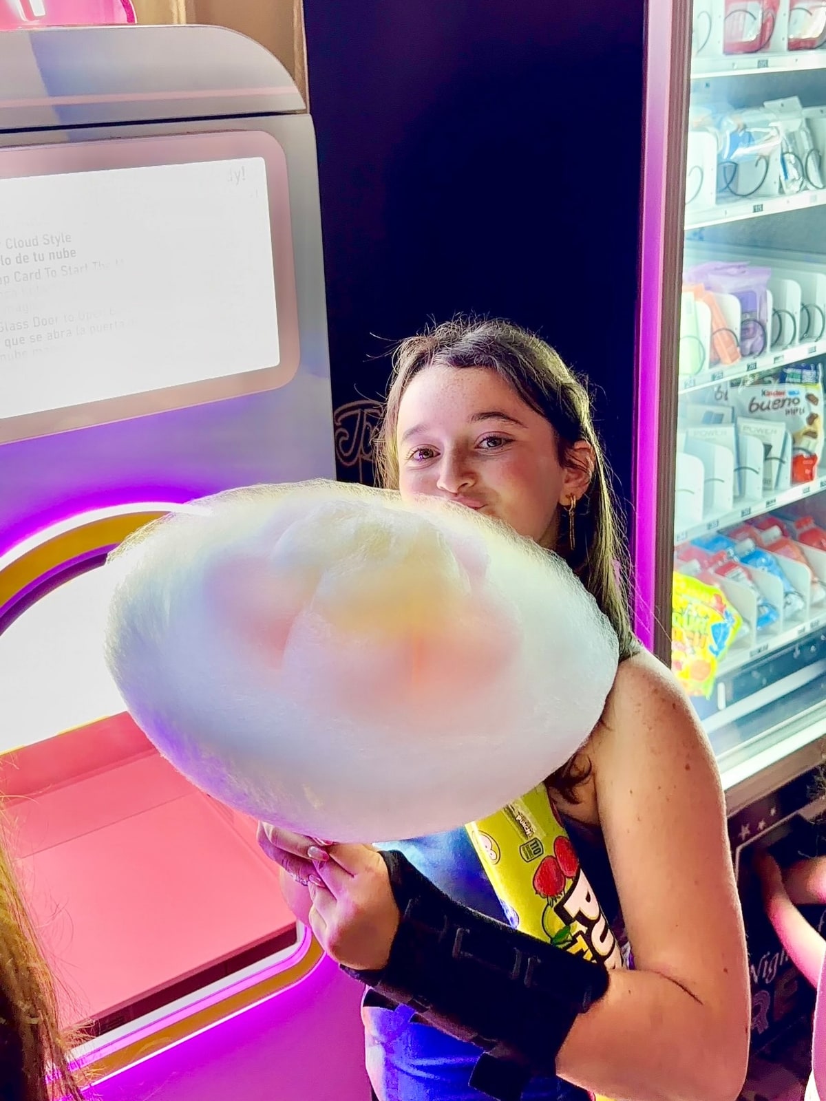 A woman holding a large pastel-colored cotton candy beside a vending machine.
